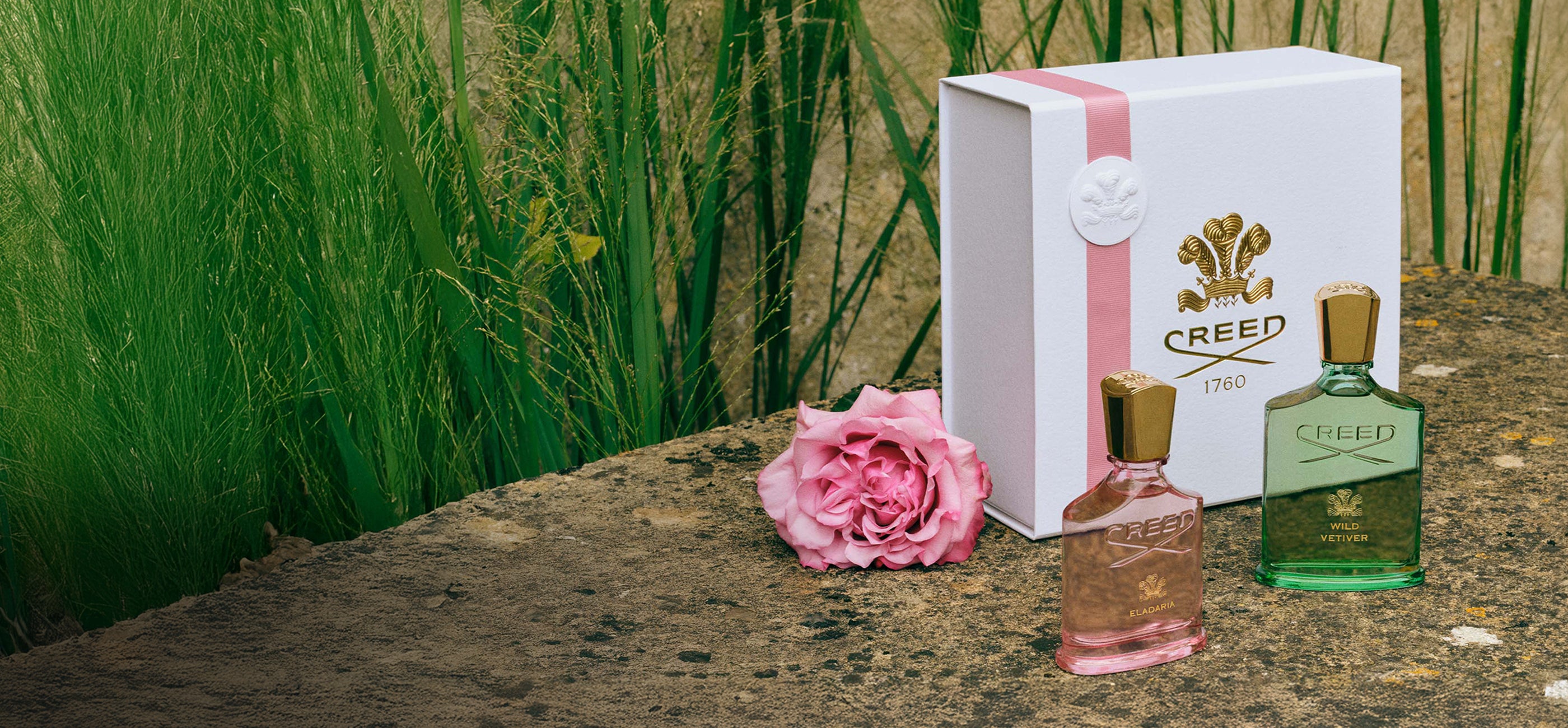 Two Creed perfume bottles and their packaging on a natural stone surface with grass in the background.